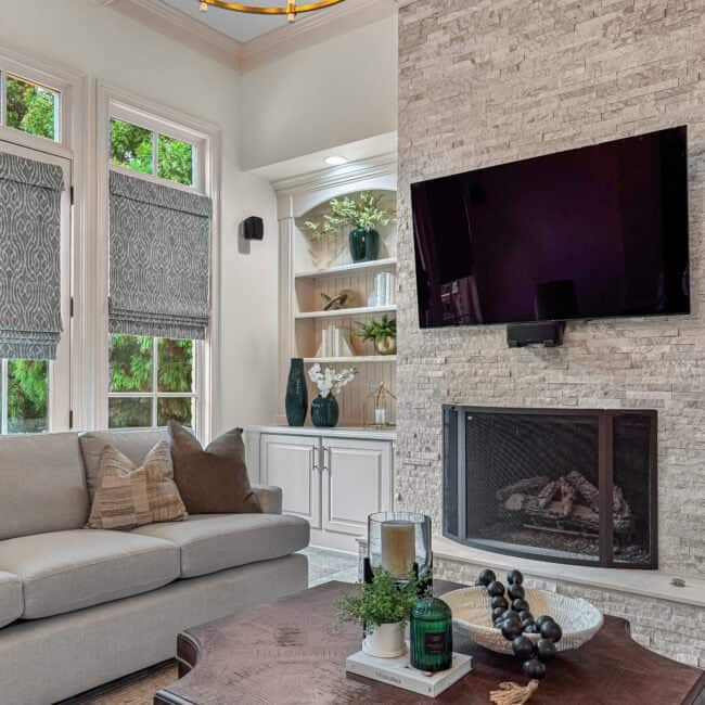 Bright and inviting living room featuring a stone fireplace, wall-mounted TV, and natural light through large windows.