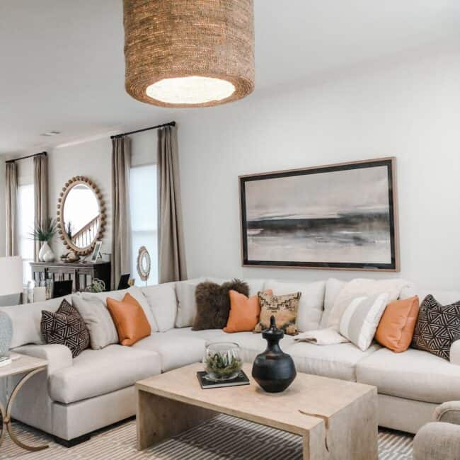 Bright living room featuring a large white sectional sofa with orange, beige, and patterned throw pillows, a beige textured area rug, and natural light from large windows.