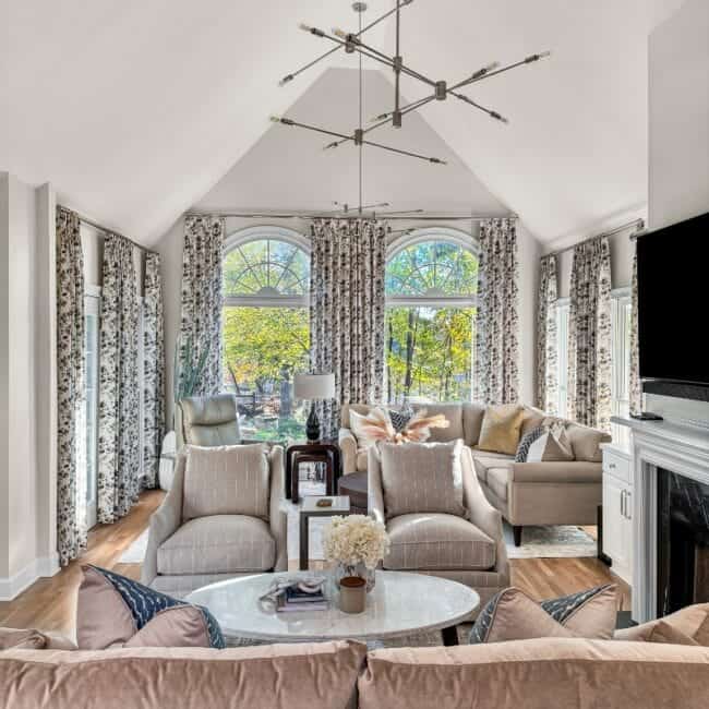 Bright, elegant living room with large windows and floral curtains. Comfortable seating and modern lighting design.