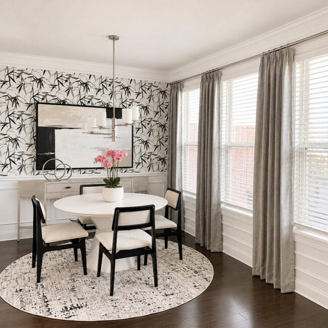 Bright dining room with round table, black chairs, floral wallpaper, and large windows.