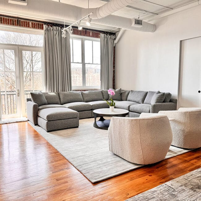 Bright living room with large windows, gray sectional sofa, and wooden floors.