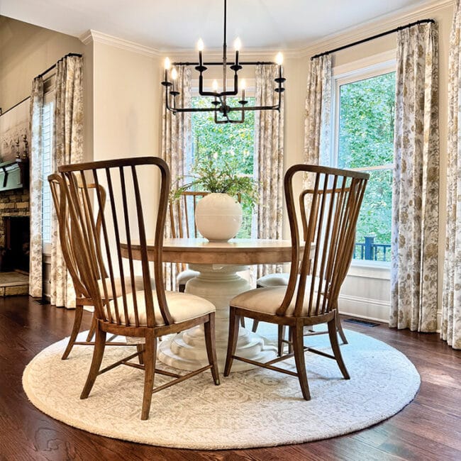 Bright dining room with round table, four wooden chairs, and large windows with floral curtains.